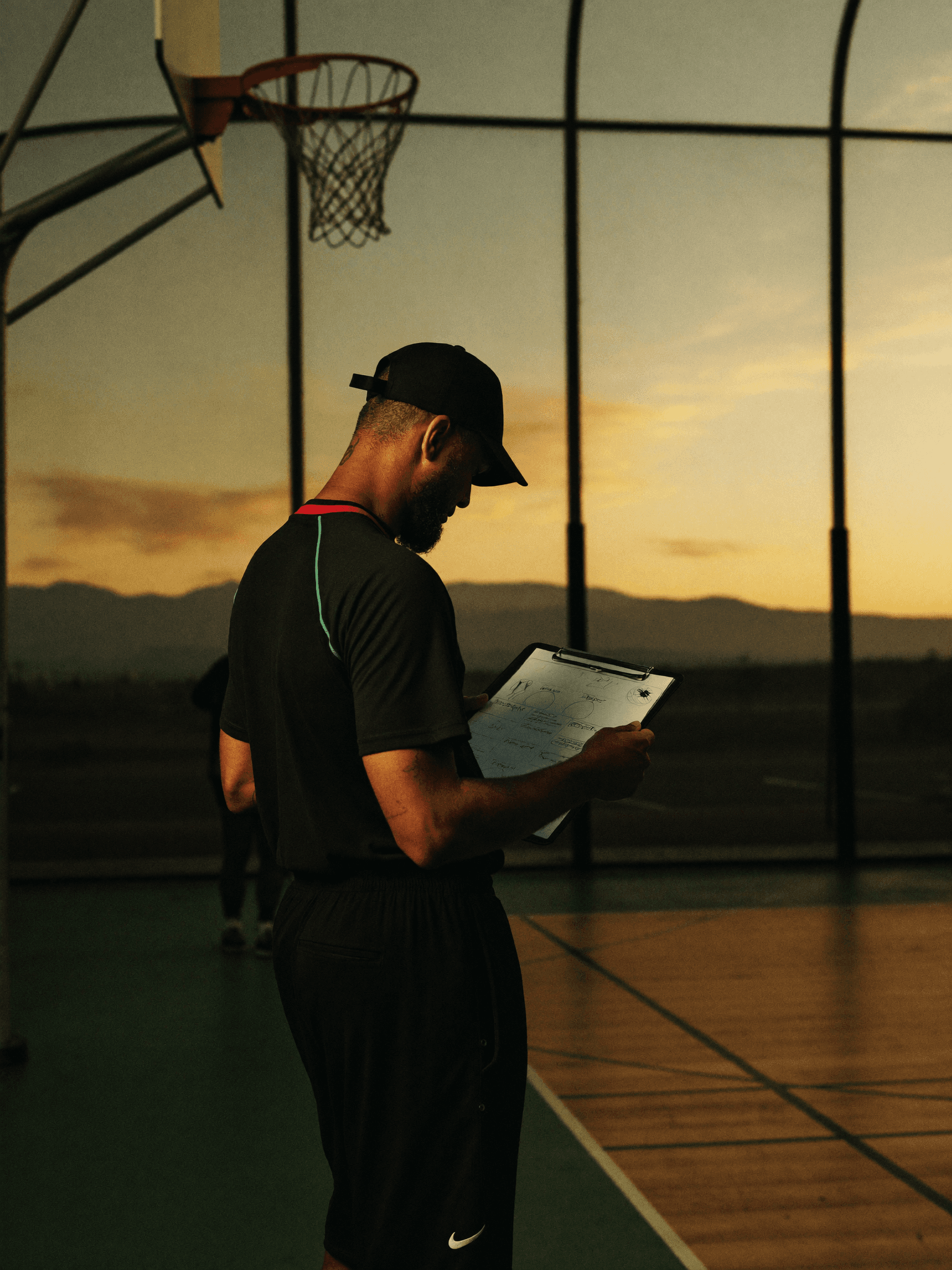Coach standing on the sideline of a gym at dusk, evaluating athletes